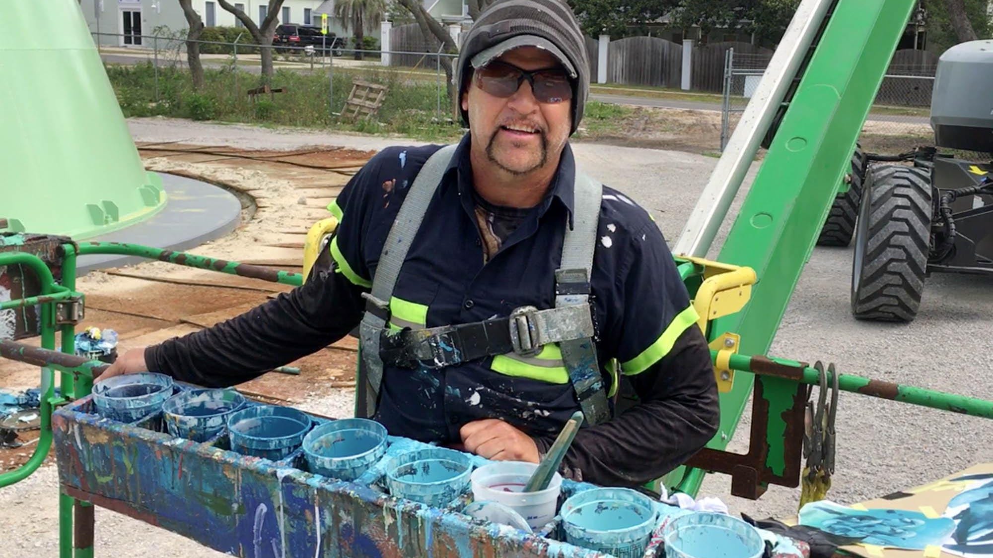 Artist Eric Henn poses with some of his paints in the bucket lift that he is using to paint sea life murals on the new water tower near Calhoun Avenue in Destin.